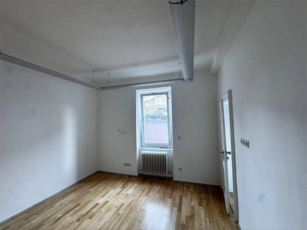 Leerer Raum mit Parkettboden, weißen Wänden, einem Fenster mit Heizkörper und sichtbaren Leitungen an der Decke.