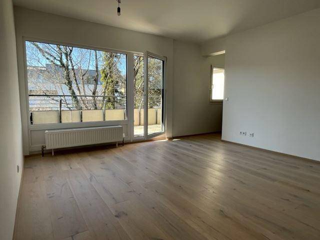 Helles, unmöbliertes Zimmer mit großen Fenstern, Holzboden und Zugang zum Balkon.