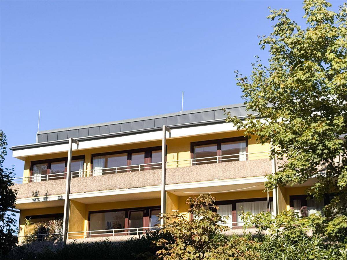 Fassade eines Mehrfamilienhauses mit Balkonen und grüner Umgebung unter blauem Himmel.