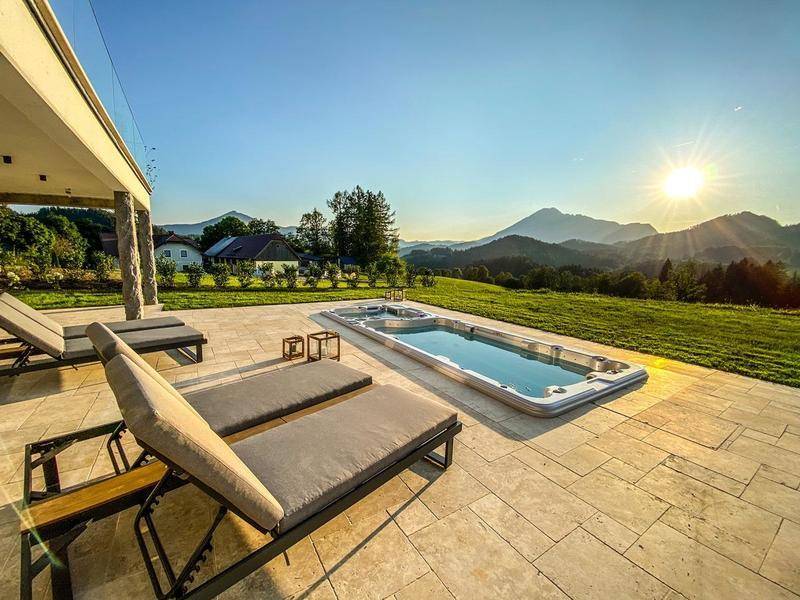 Sonnige Terrasse mit Liegestühlen, Swim-Spa und weitem Bergblick vor moderner Architektur.