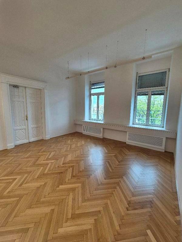 Heller Raum mit Fischgrätparkett, hohen Decken und zwei großen Fenstern, ideal als Büro.