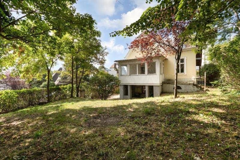 Rückseite des Hauses mit großem Gartenbereich, einer verglasten Veranda und vielen Bäumen im Hintergrund.