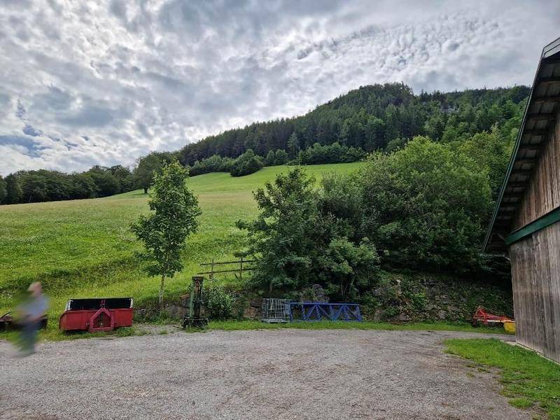 Außenansicht eines landwirtschaftlichen Gebäudes mit Schotterfläche und grünen Hügeln im Hintergrund.