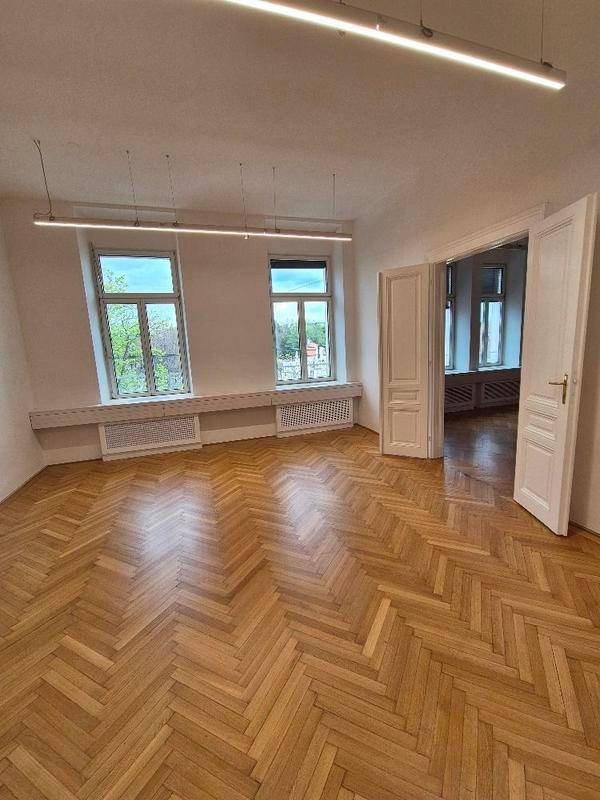 Geräumiger Büroraum mit Fischgrätparkett, viel Tageslicht und Zugang zu einem weiteren Zimmer.
