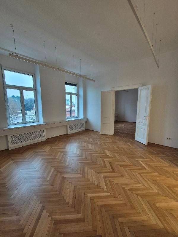 Heller Büroraum mit Fischgrätparkett, zwei großen Fenstern und moderner Deckenbeleuchtung.