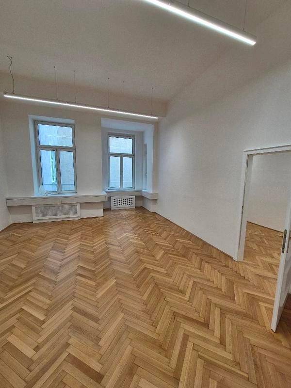 Heller Büroraum mit Fischgrätparkett, zwei großen Fenstern und moderner Deckenbeleuchtung.