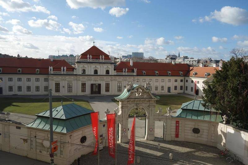 Panoramablick auf ein historisches Gebäude mit weitläufigem Hof und roter Dachlandschaft unter blauem Himmel.