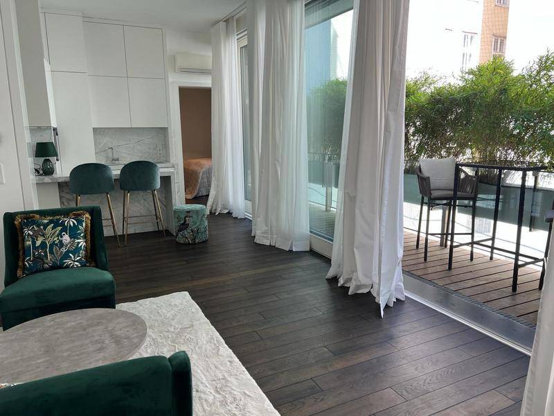 Helles Wohnzimmer mit dunklem Holzboden, grünen Samtsesseln und Zugang zu einem möblierten Balkon.
