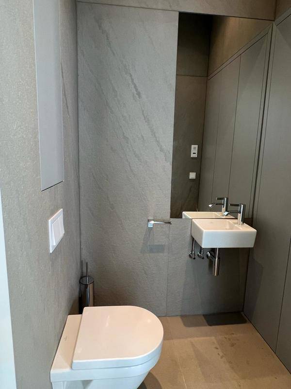 Kompaktes Gäste-WC mit grauen Fliesen, wandhängender Toilette und kleinem Waschbecken mit Spiegel.