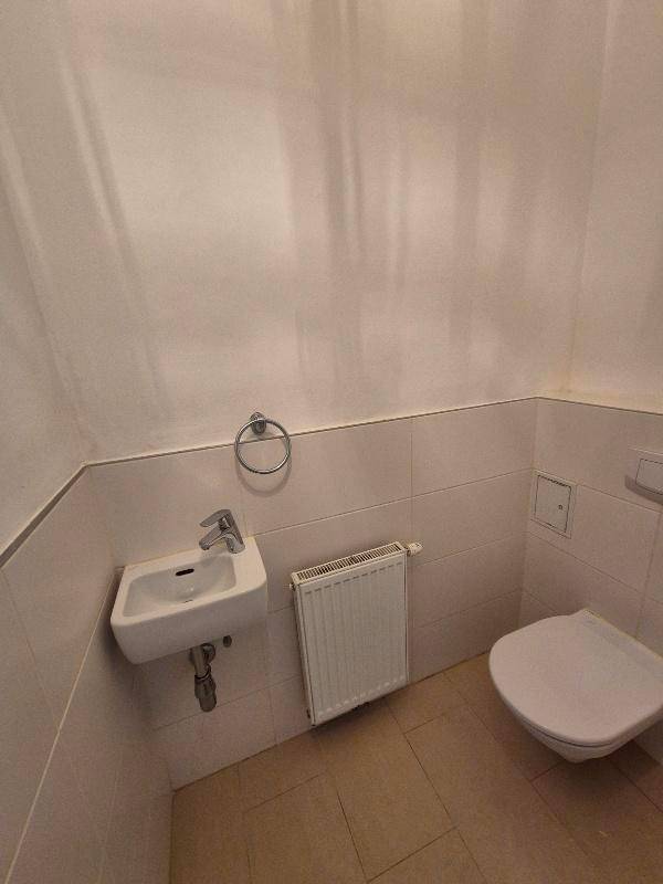 Separates WC mit Wand-WC, kleinem Waschbecken und Heizkörper, ausgestattet mit hellen Fliesen.