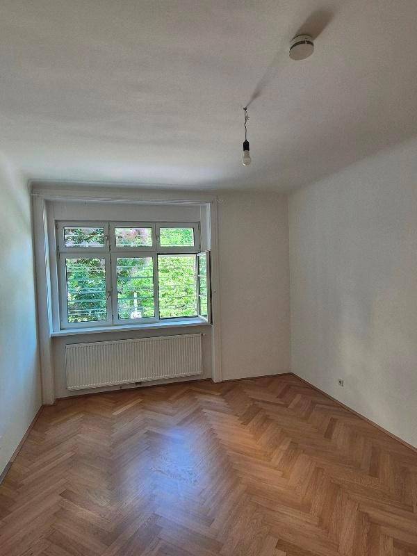 Geräumiges, leeres Zimmer mit Fischgrätparkett und großem Fenster, ideal als Schlaf- oder Arbeitszimmer.