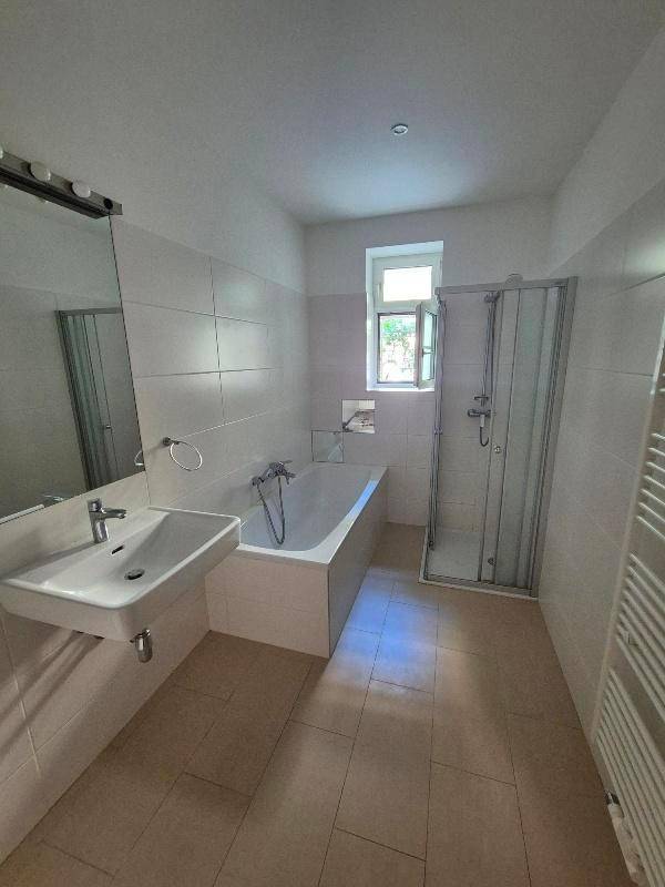 Helles Badezimmer mit Badewanne, Dusche, Waschbecken und Fenster, ausgestattet mit hellen Fliesen.