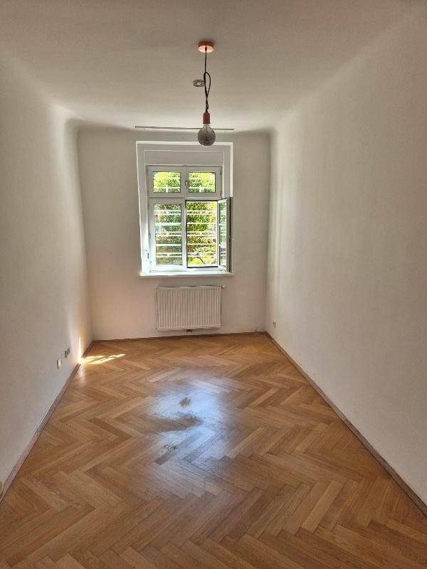 Helles, leeres Zimmer mit Fischgrätparkett und einem Fenster, das viel Tageslicht hereinlässt.