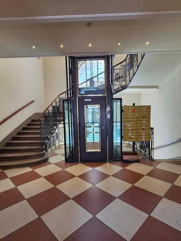Eingangsbereich mit Schachbrettmuster-Boden, Treppe und modernem Glasaufzug.