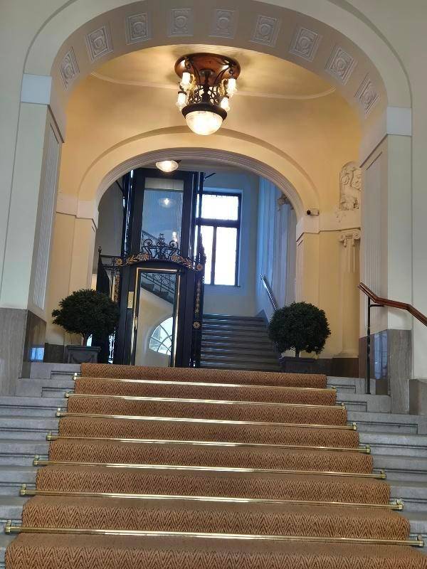 Prunkvoller Eingangsbereich mit breiter Treppe, Teppichläufer und einem historischen Aufzug in einem Altbau.