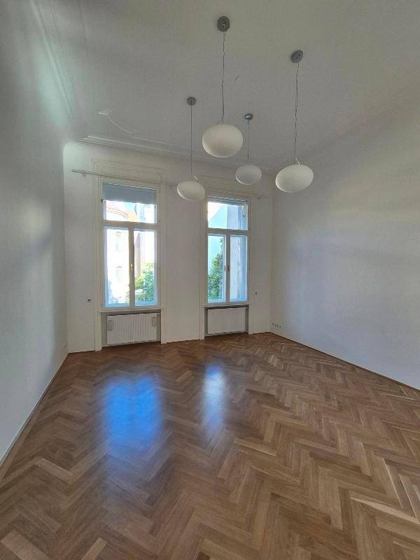 Geräumiges Zimmer mit glänzendem Fischgrätparkett, hohen Fenstern und modernen Hängelampen.