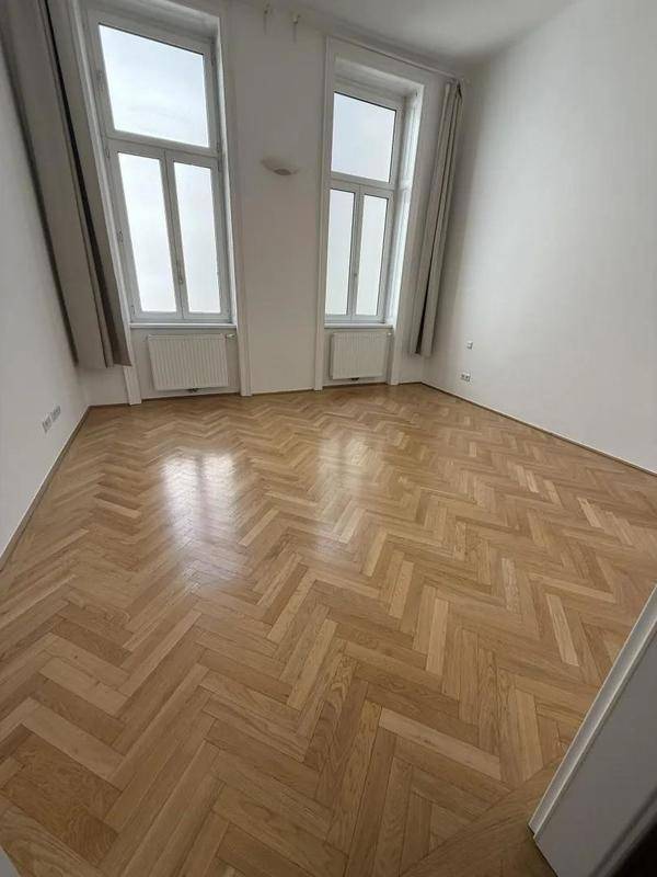 Leeres Zimmer mit klassischem Fischgrätparkett, weißen Wänden und hohen Fenstern.