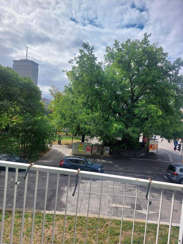 Blick vom Balkon auf eine belebte Straße mit Bäumen und Gebäuden im Hintergrund unter bewölktem Himmel.