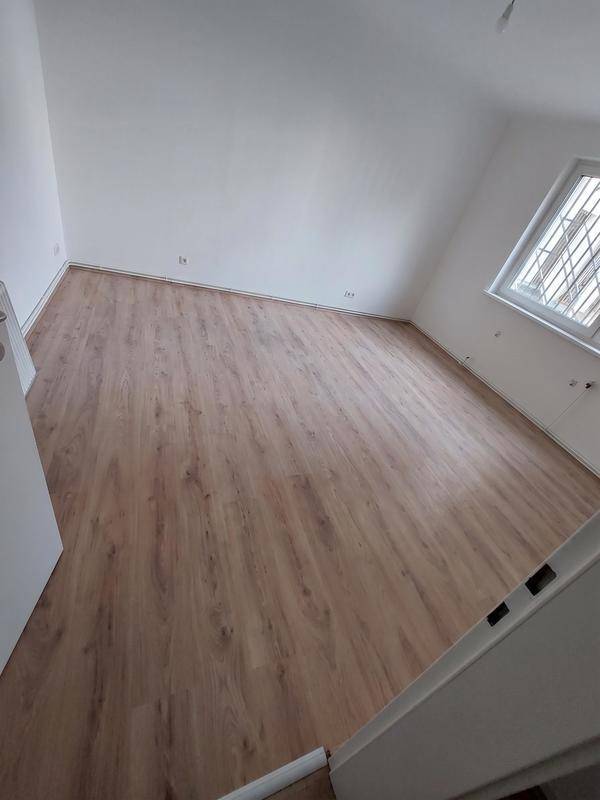 Helles, leeres Zimmer mit Laminatboden, weißen Wänden und einem Fenster mit Gitterstäben.