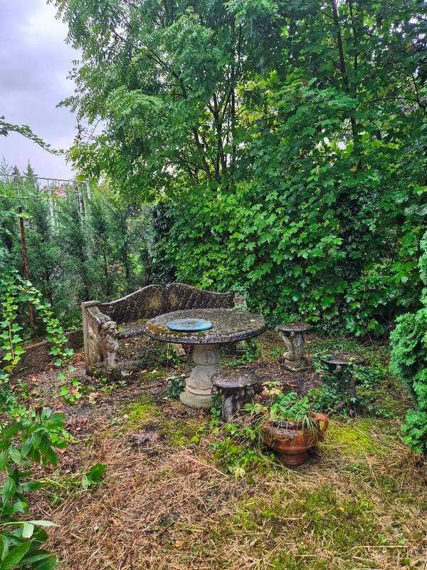 Verwilderter Gartenbereich mit rustikalen Steinmöbeln, umgeben von dichtem Grün und Bäumen.