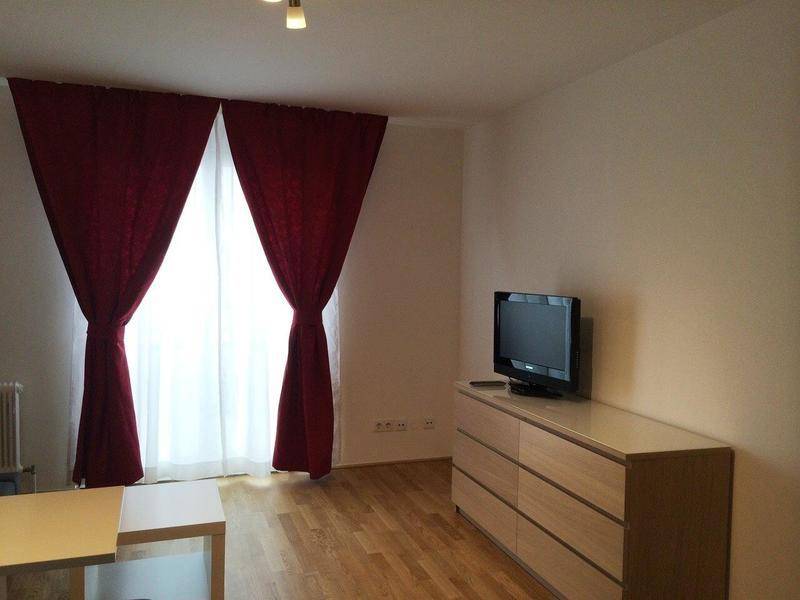 Wohnbereich mit Fernseher auf einer Kommode, Parkettboden und roten Vorhängen am Fenster.