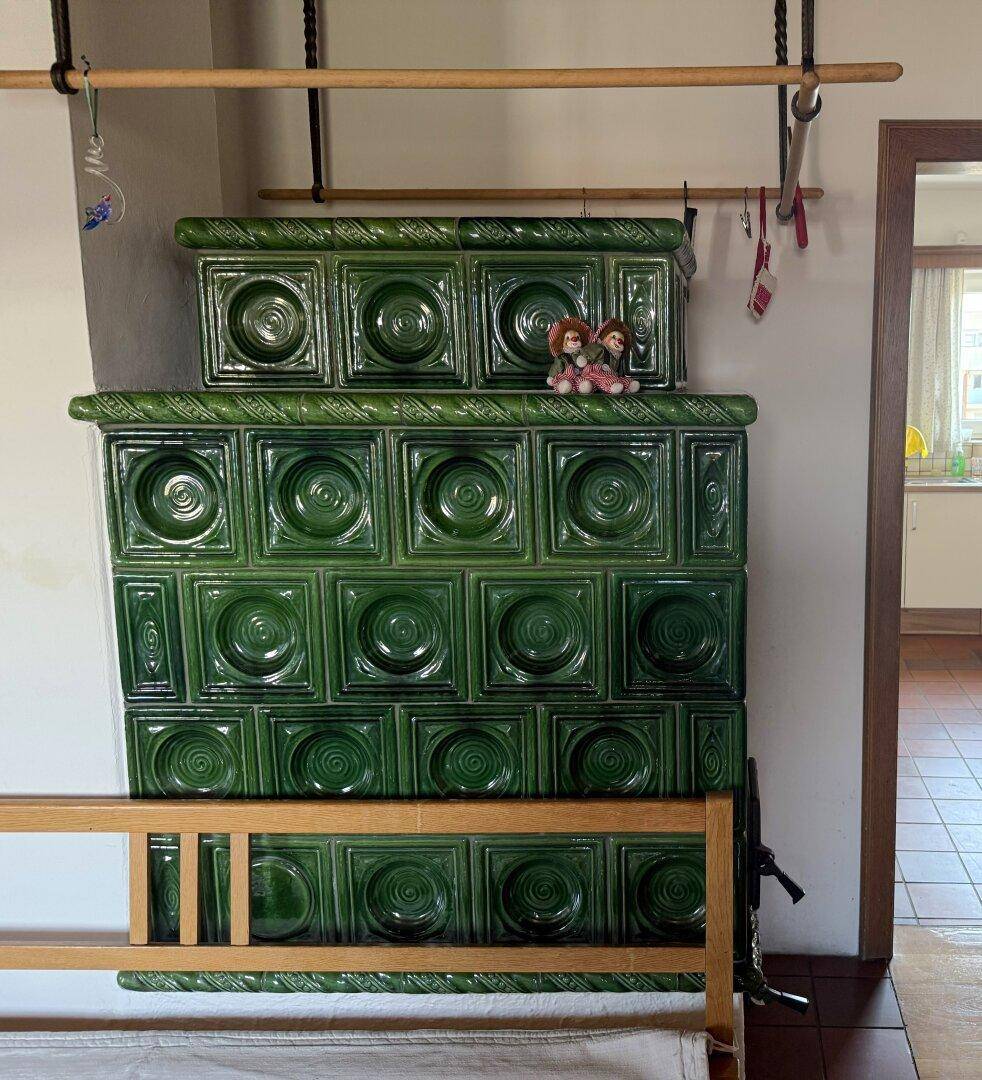 Grüner Kachelofen als traditionelle Heizquelle, verziert mit Puppen, in einem Innenraum.