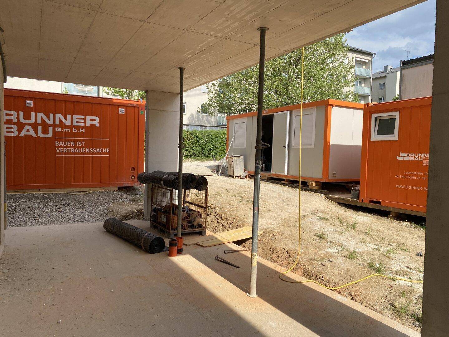 Baustelle im Erdgeschoss mit Betonstützen und orangefarbenen Baucontainern im Hintergrund.
