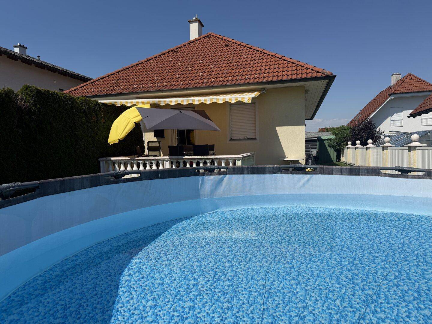 Hausansicht mit Terrasse und einem oberirdischen Pool im Vordergrund unter blauem Himmel.