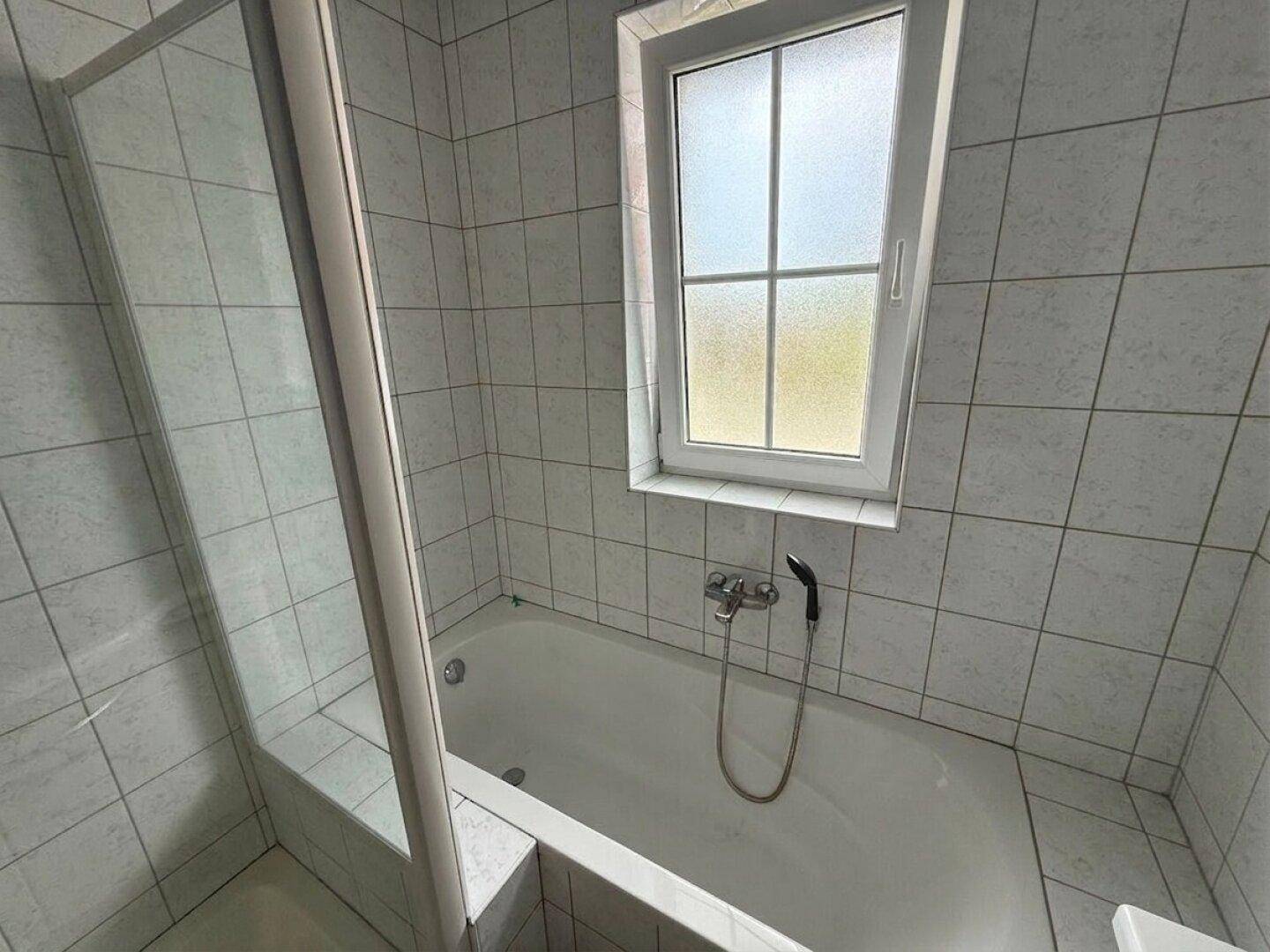Badezimmer mit Badewanne, Duschabtrennung und Fenster, komplett gefliest in hellem Design.