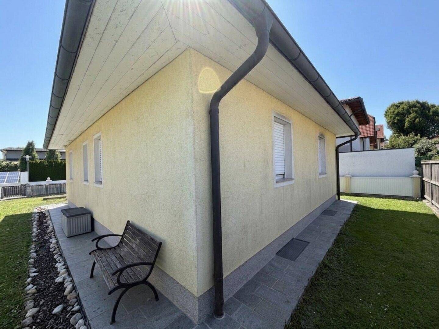 Eckansicht des Hauses mit Fenstern, Rollläden und einer kleinen Holzbank im Gartenbereich.