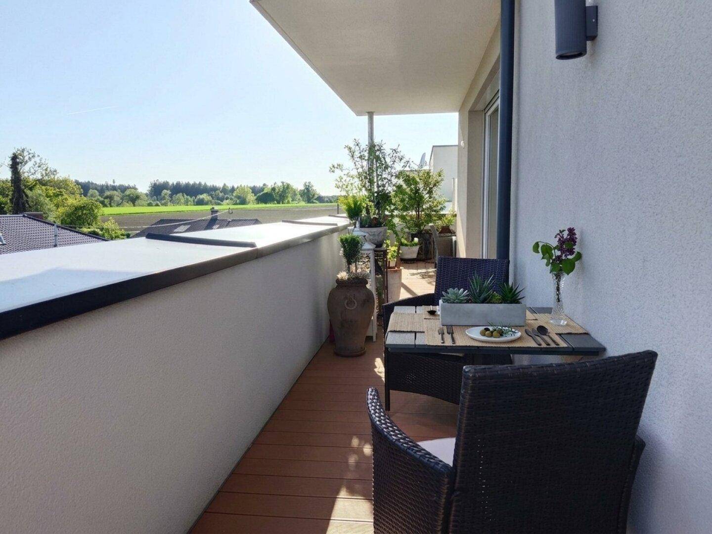 Sonniger Balkon mit kleinem Esstisch, Pflanzen und Blick auf grüne Felder und Häuser.