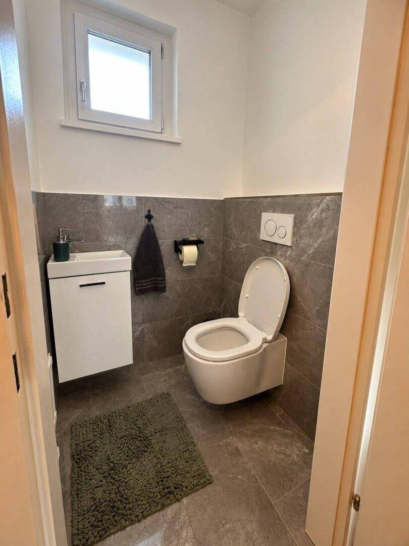 Modernes Gäste-WC mit wandhängender Toilette, kleinem Waschbecken und grauen Fliesen.