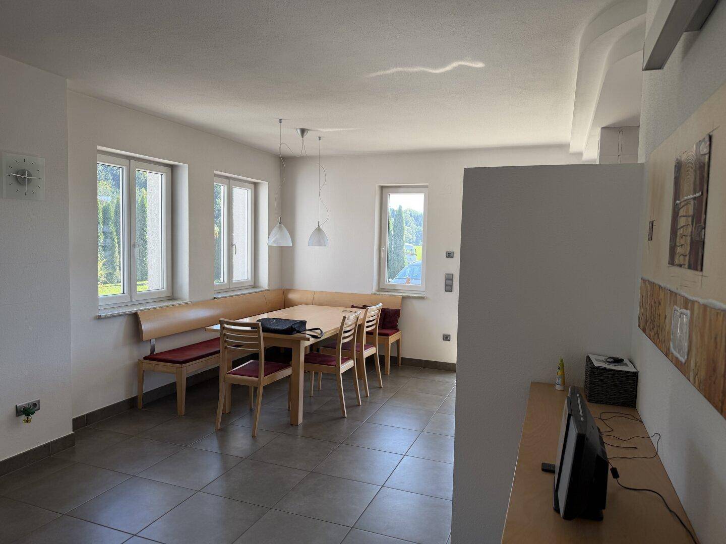 Heller Essbereich mit großer Eckbank, Holztisch und Fliesenboden, umgeben von Fenstern.