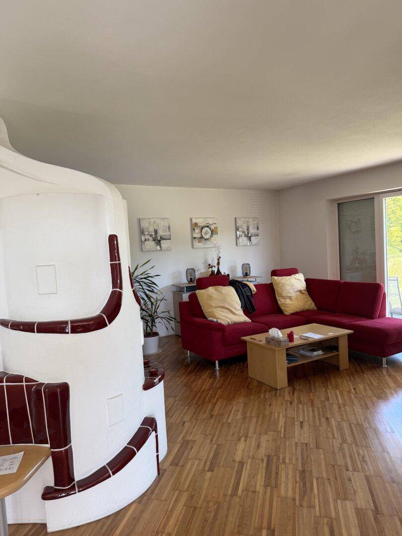 Gemütliches Wohnzimmer mit rotem Ecksofa, Parkettboden und einem traditionellen Kachelofen.