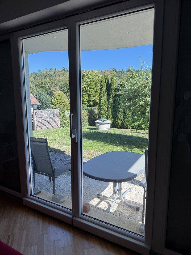 Blick vom Wohnzimmer durch die große Glastür auf den grünen Garten mit Sitzbereich.