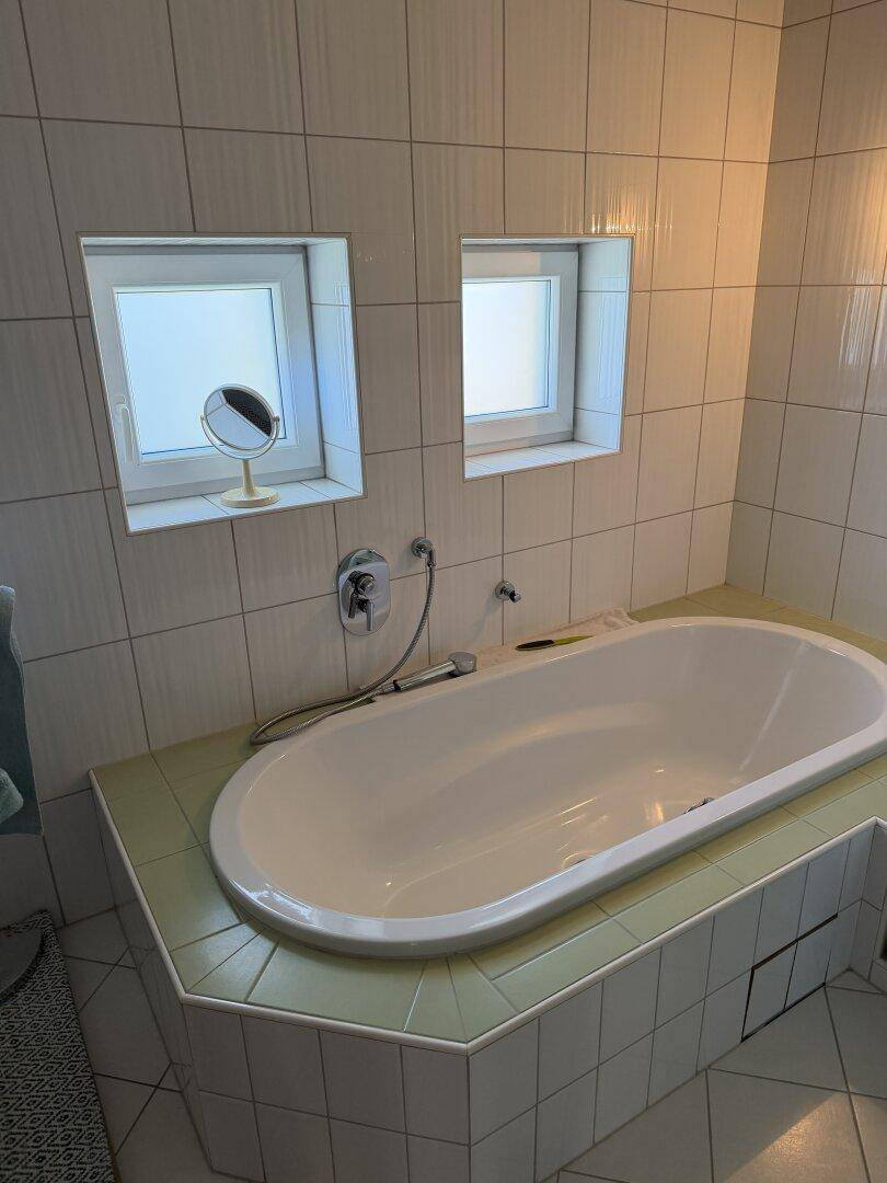 Badezimmer mit eingebauter Badewanne und hellgrünen Fliesen, ausgestattet mit zwei Fenstern.