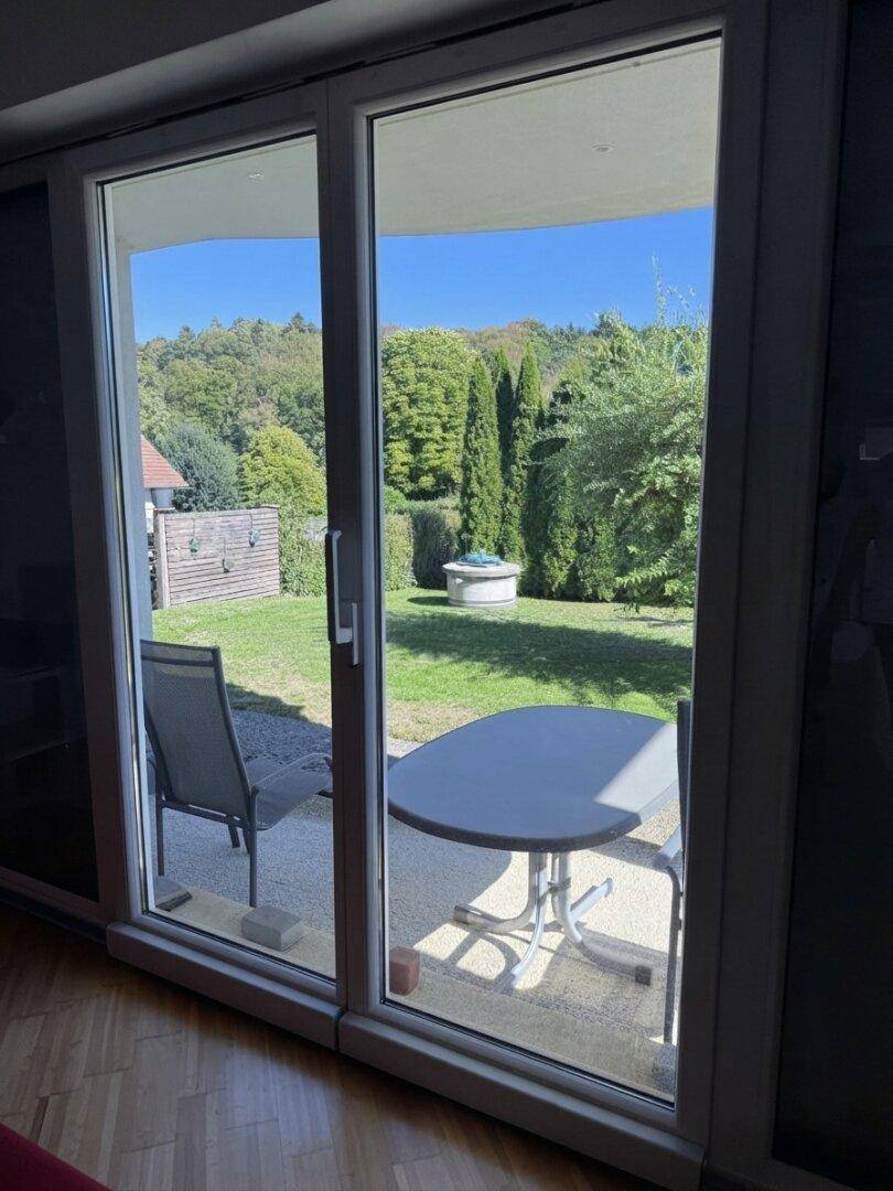 Blick durch eine große Glastür auf eine möblierte Terrasse und den grünen Gartenbereich.