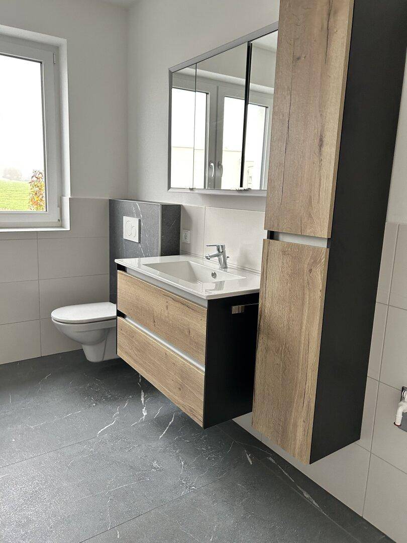 Modernes Badezimmer mit schwebendem Waschtisch, WC und Fenster, ausgestattet mit dunklen Bodenfliesen.
