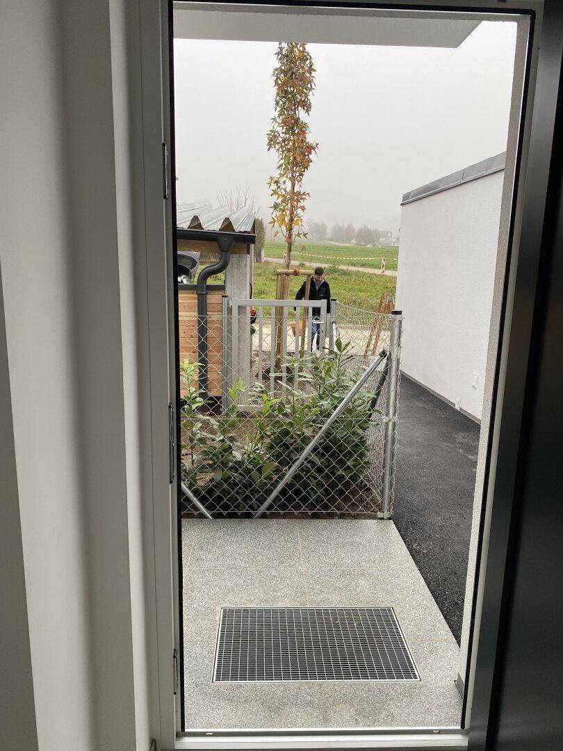 Blick von der Eingangstür auf einen gepflasterten Bereich mit Zaun und dahinterliegendem Feld an einem bewölkten Tag.