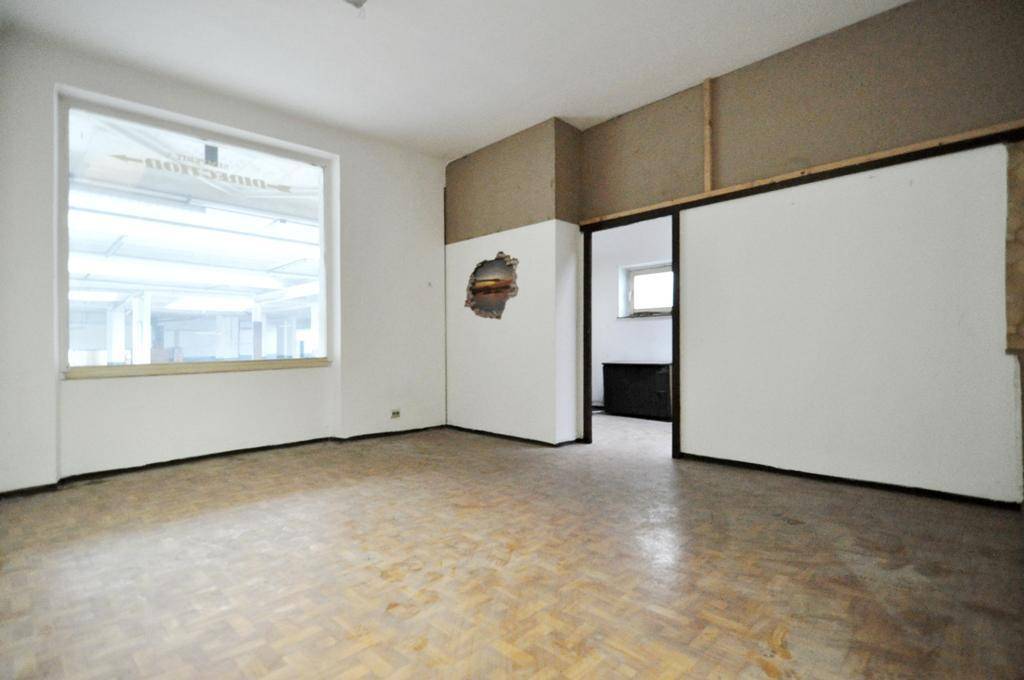 Leeres Büro mit Fischgrätparkett, großem Fenster und Durchgang zu einem weiteren Raum, der renovierungsbedürftig ist.