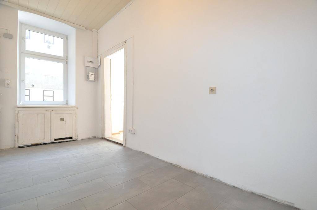 Leerer Raum mit grauen Fliesen, weiß gestrichenen Wänden und einem Fenster mit Heizungsverkleidung.
