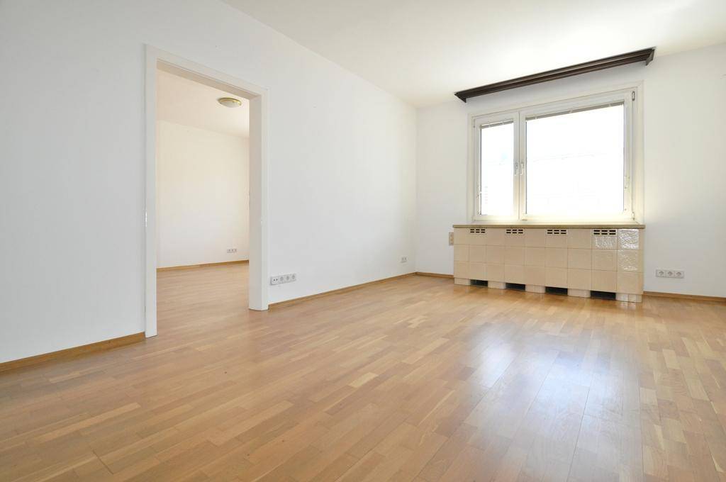 Heller Wohnraum mit Parkettboden, Fenster und einem Durchgang zu einem weiteren Zimmer.