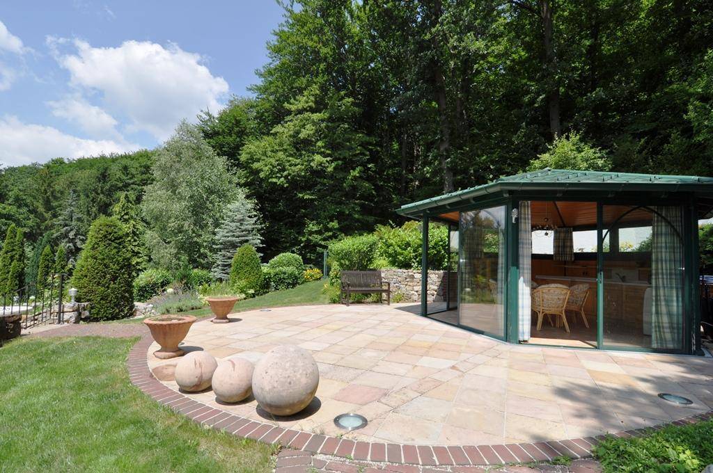 Geräumige Steinterrasse mit einem verglasten Gartenpavillon, umgeben von üppiger Natur und Bäumen.