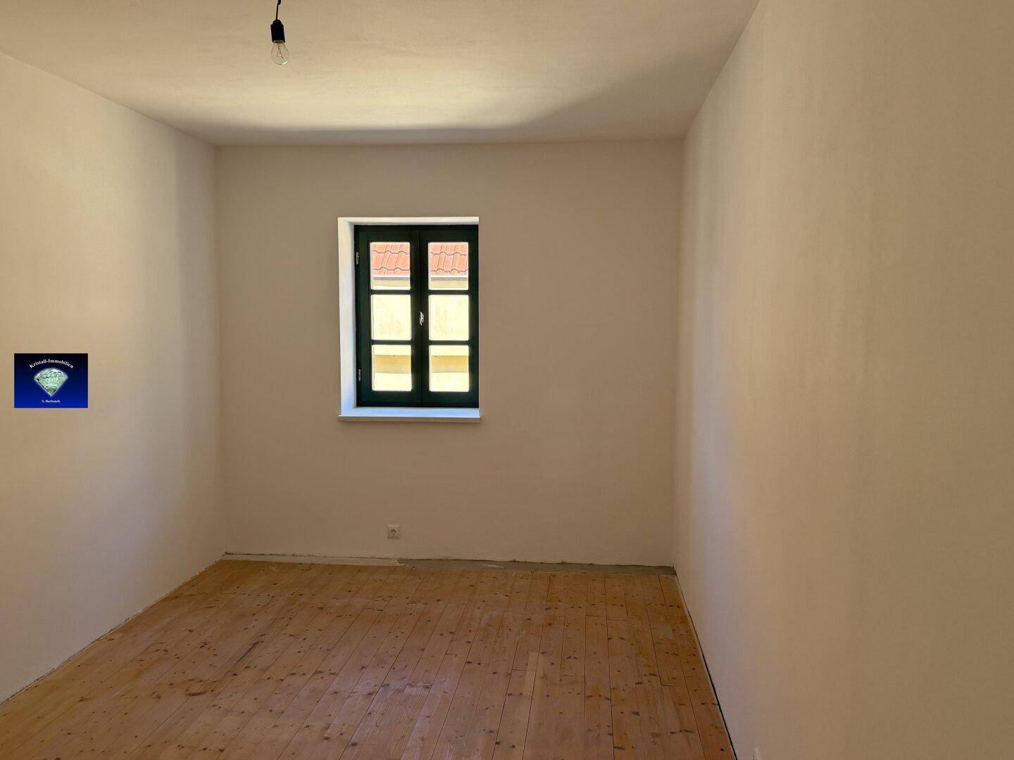 Heller Raum mit Holzdielenboden, weißen Wänden und einem grünen Fenster, ideal für ein Schlafzimmer.