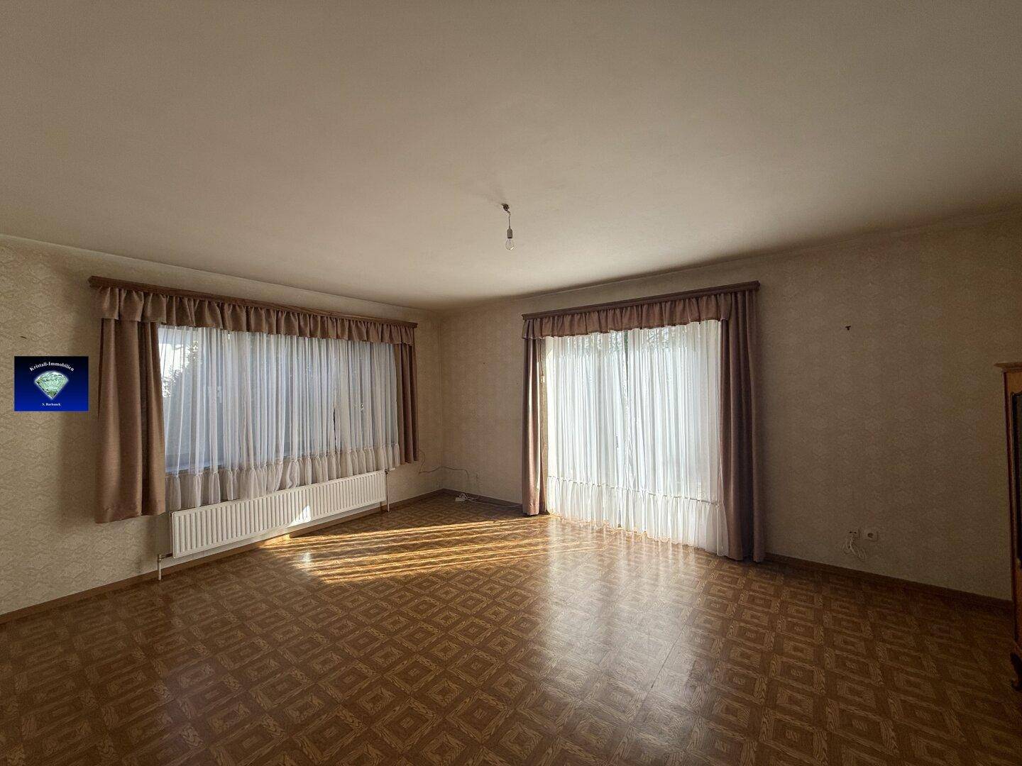 Helles Wohnzimmer mit zwei großen Fenstern, Heizkörpern und gemustertem Bodenbelag.