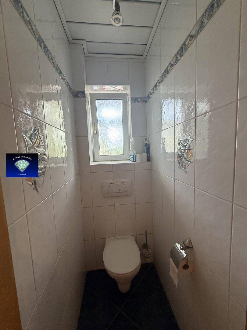 Kleines WC mit Toilette, Fenster und hellen Fliesen an Wänden und Boden.