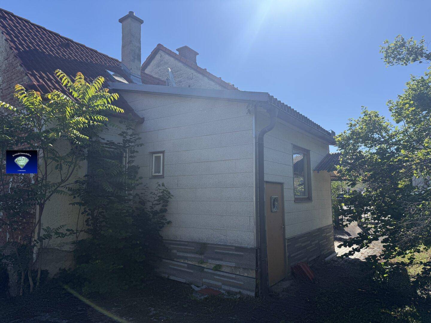 Seitenansicht des Hauses mit heller Fassade, kleinem Fenster und Nebeneingang.