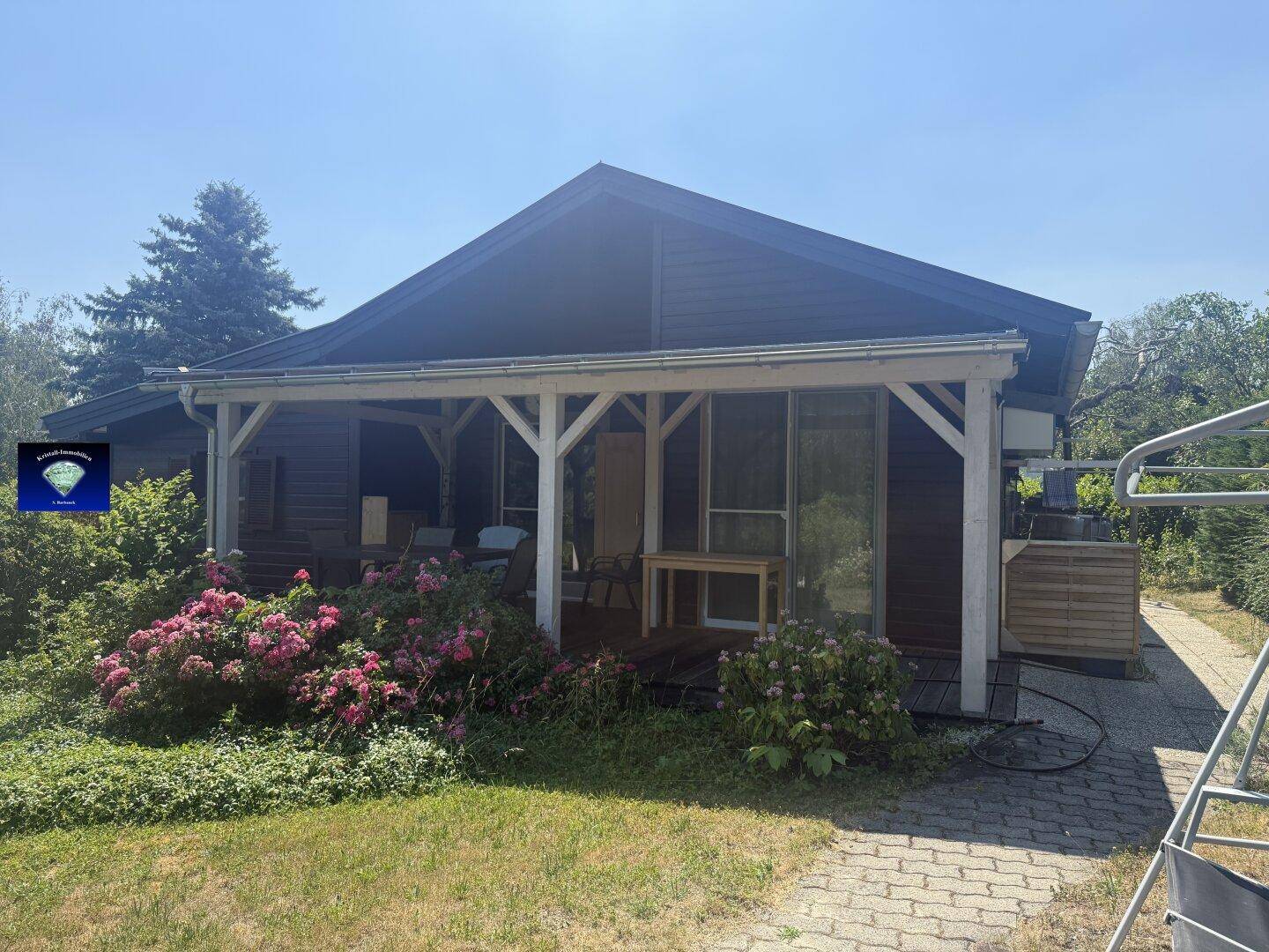Überdachte Terrasse mit Gartenmöbeln und blühenden Sträuchern vor dem Haus.