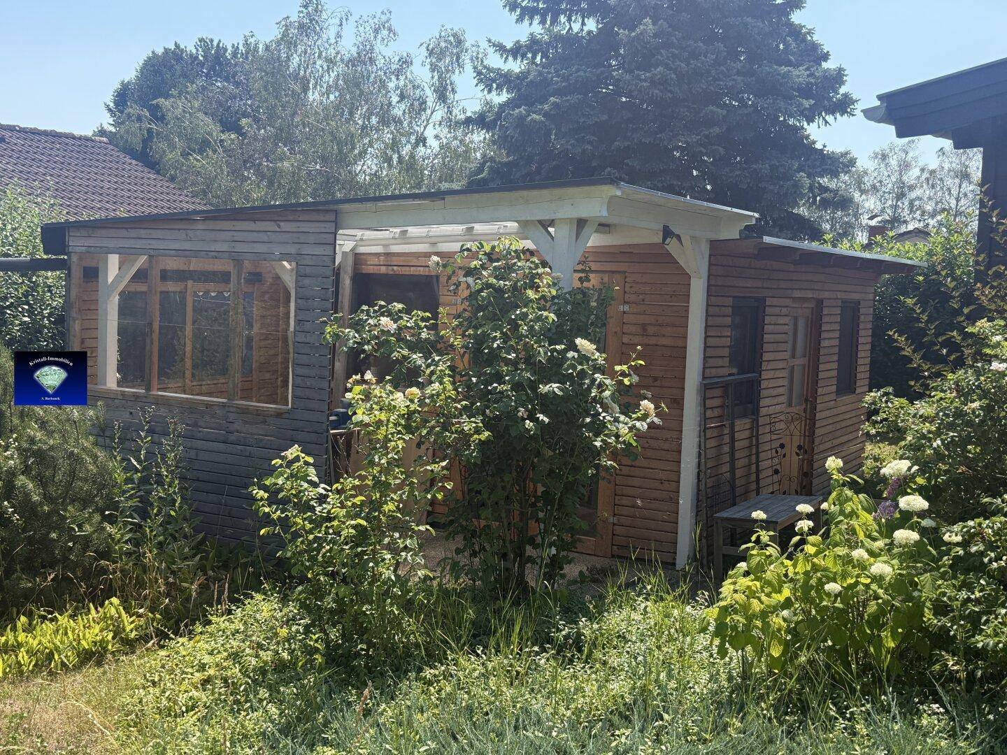 Kleines Holzhaus im Garten mit überdachtem Bereich, umgeben von üppigem Grün.