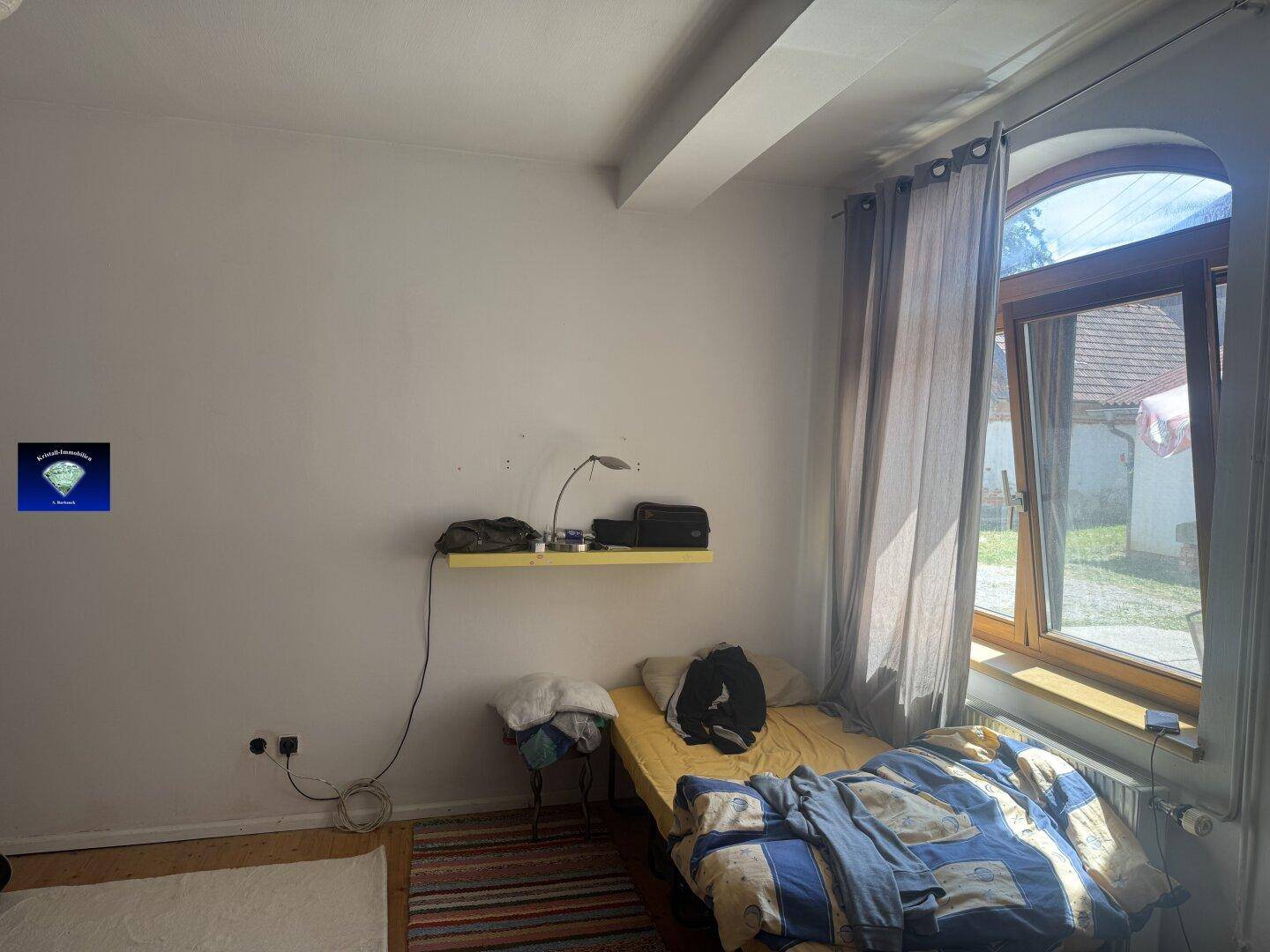 Helles Schlafzimmer mit einem Bett, einem Regal und einem großen Bogenfenster mit Blick ins Grüne.
